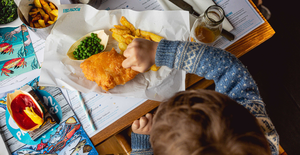 Child eating fish and chips with peas at a restaurant table.